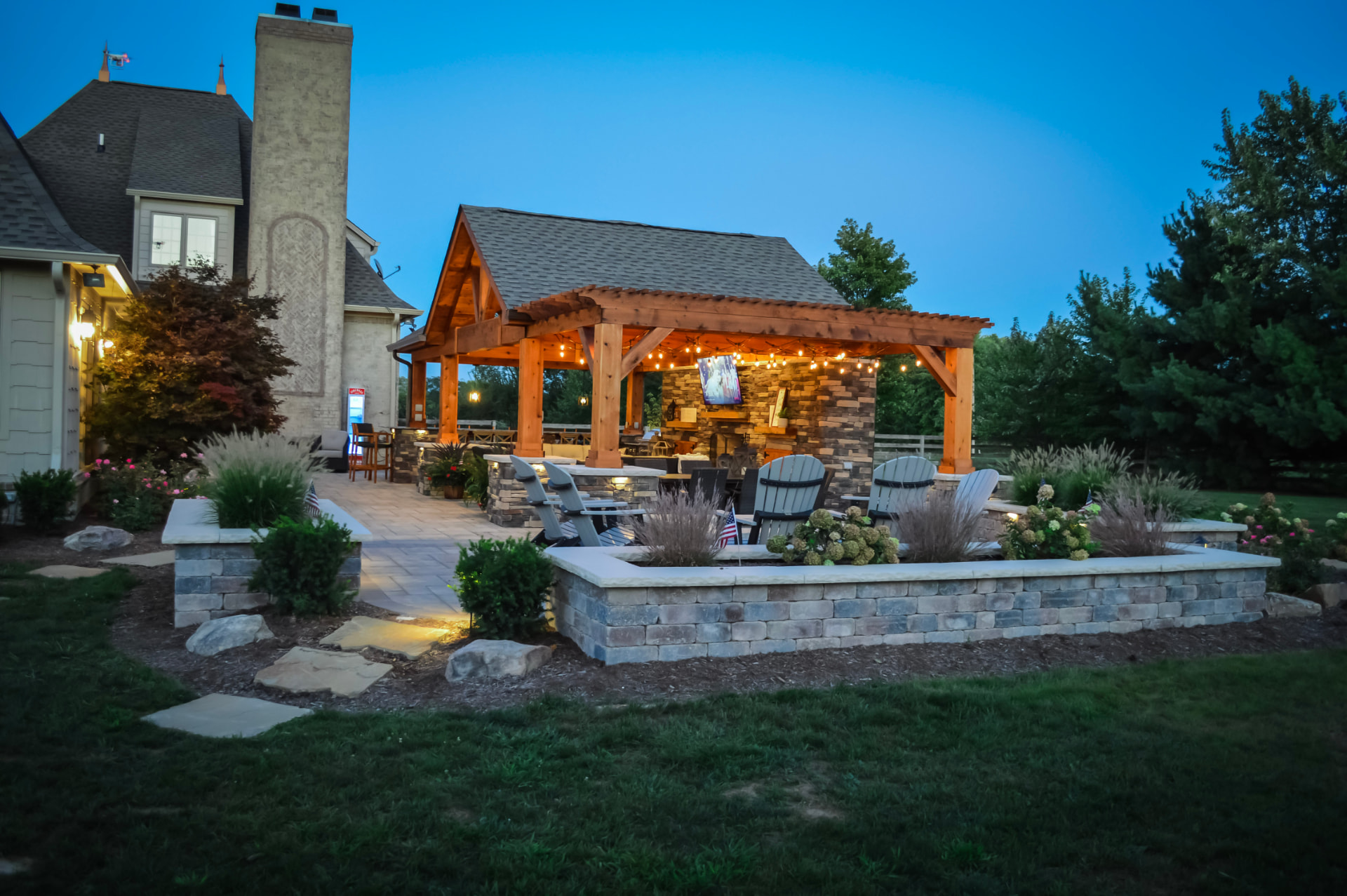 Custom outdoor living space with a timber pergola, stone patio, fire feature, landscape lighting and planted garden beds at dusk